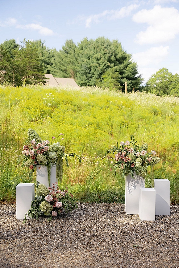 Ceremony altar decor with outdoor wedding altar plinths topped with roses, hydrangea and greenery in a wildflower meadow under blue sky