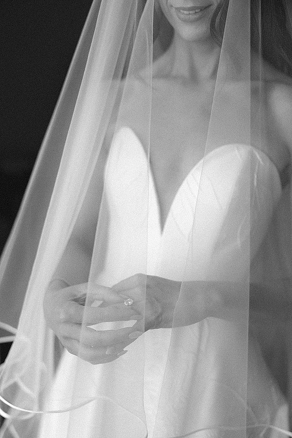 Bridal portrait of a bride under veil in a strapless wedding dress, hands clasped showing an engagement ring against a dark interior background