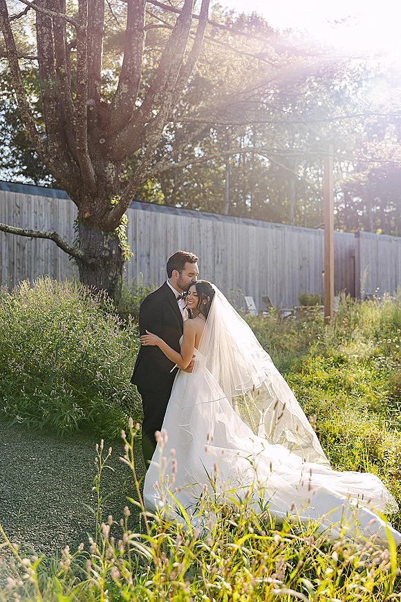 Couple portrait of bride and groom hug with a forehead kiss, cathedral veil flowing in sunlit garden by a rustic fence