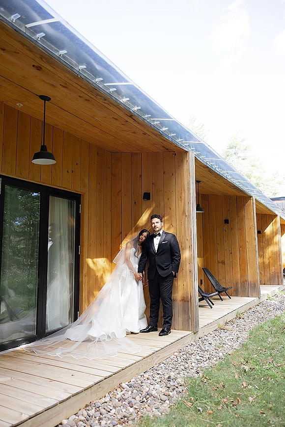 Couple portrait of bride in strapless wedding dress and long veil leaning on groom in black tuxedo by a wood cabin porch deck in daylight