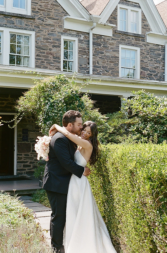 Couple portrait of bride and groom hug as she holds a wedding bouquet, standing by a stone house walkway with hedges and greenery