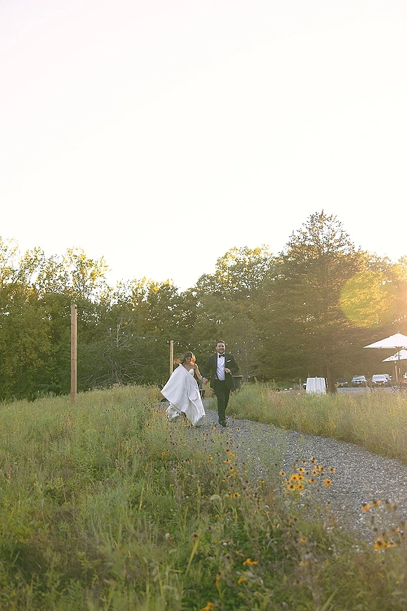 Wedding couple portrait of bride and groom walking hand in hand down a gravel path through wildflowers under string lights at sunset, bow tie and strapless dress visible