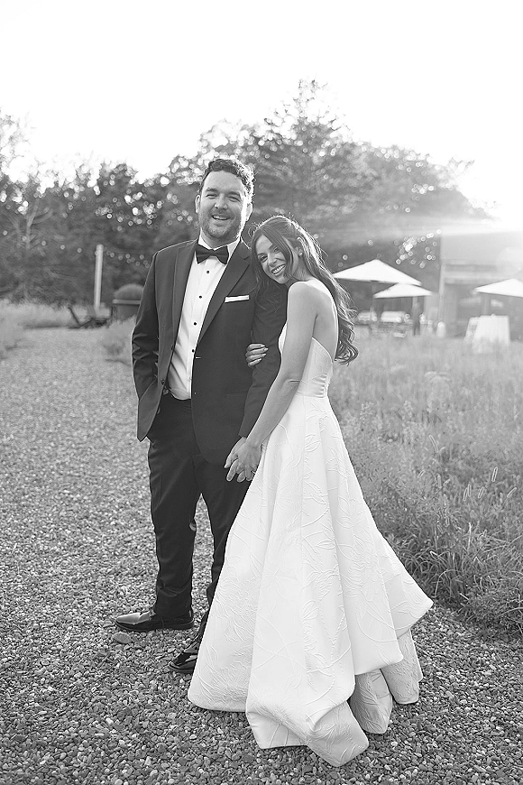 Couple portrait of bride in strapless wedding dress leaning on groom in tuxedo, holding hands with rings on a gravel path at golden hour