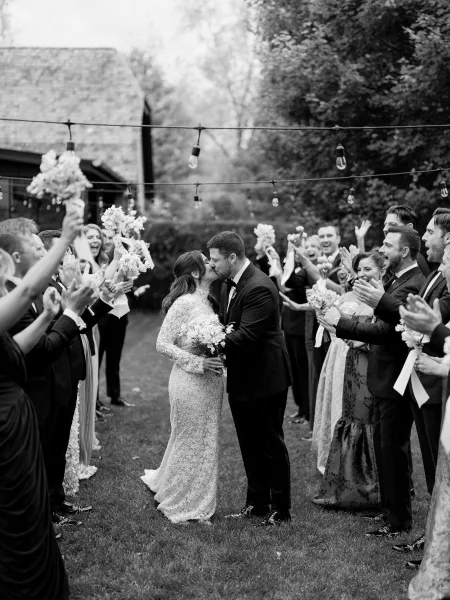 Ceremony exit kiss as newlyweds make their wedding recessional, bouquet ribbons streaming beside a rustic building.