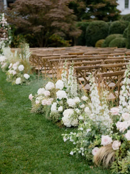 Ceremony aisle flowers create elegant outdoor ceremony decor, with blush roses edging a grass lawn amid garden hedges and trees.