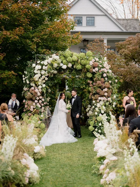 Ceremony moment as the couple stands beneath a lush floral arch during an outdoor wedding ceremony on a garden lawn beside a house.