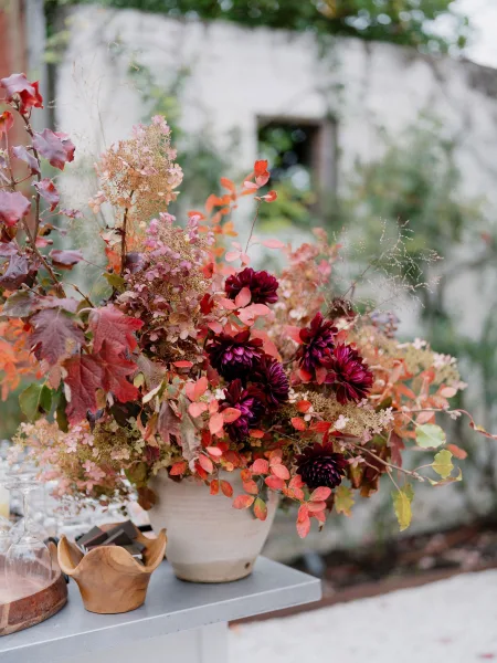 Wedding centerpiece in a fall floral arrangement featuring dahlias, framed by garden greenery, trailing vines, and a nearby stone wall.