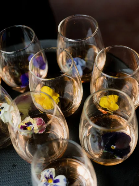 Edible flower cocktails, floral garnish drinks in stemless wine glasses on a chilled serving tray beaded with water droplets against a dark backdrop.