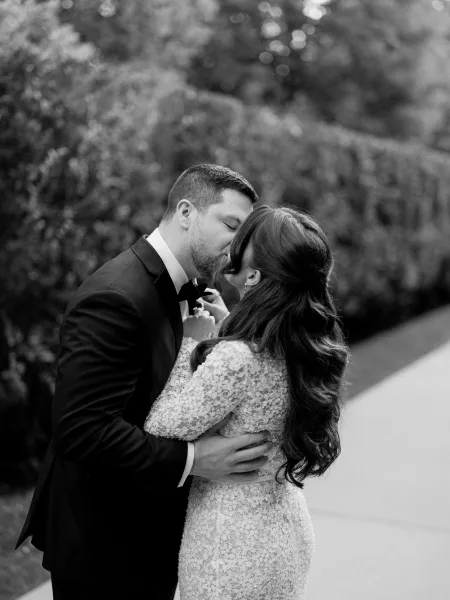 Wedding kiss portrait as a bride and groom kiss by a garden hedge, her lace bridal gown brushing the sidewalk as they embrace.