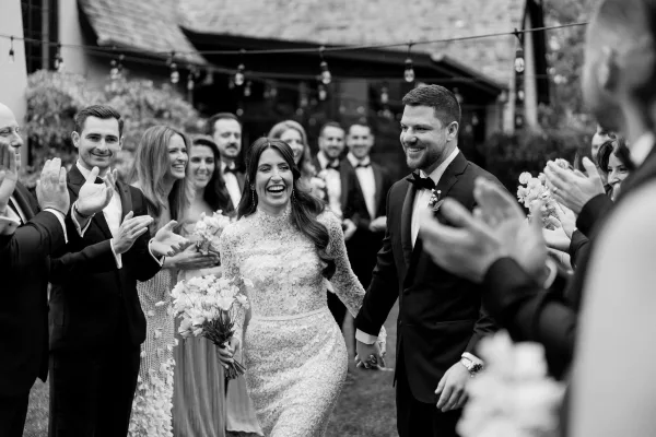 Wedding recessional during the just married exit as the bride and groom walk hand in hand on a garden lawn, her bridal bouquet under string lights.