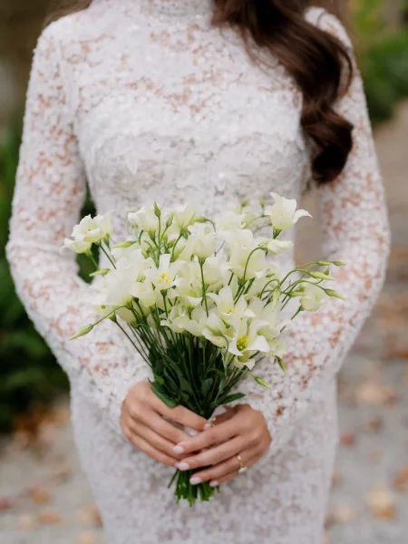 Bouquet close-up of a white bridal bouquet with soft blooms and greenery, nestled against a lace bridal gown amid garden leaves.