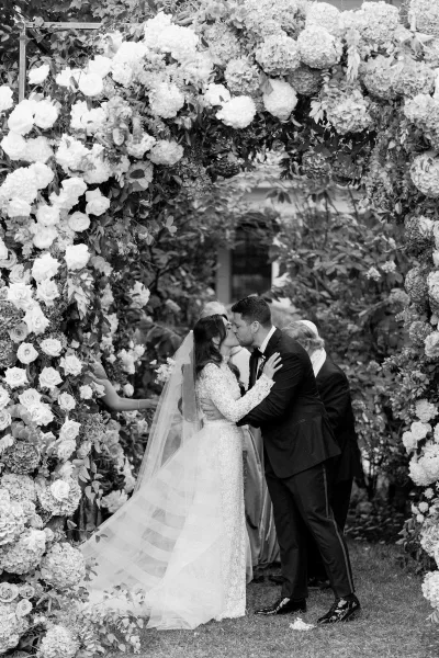 Wedding first kiss as a couple under a flower arch in a lush garden with trees and a sunlit lawn during their outdoor ceremony.