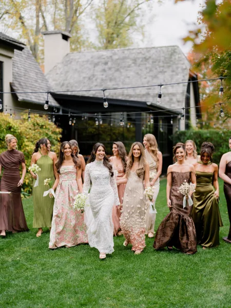 Bride and bridesmaids featuring mix and match bridesmaids stand on a garden lawn, carrying ribbon-wrapped bouquets beside hedges and a house.