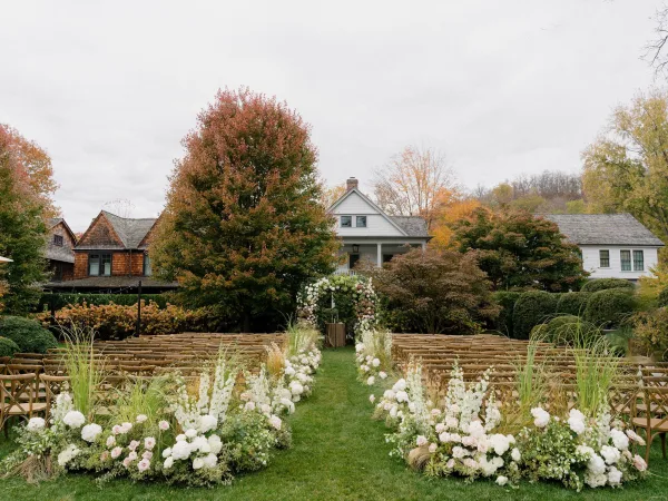 Outdoor ceremony setup with garden ceremony decor highlighted by a floral arch, backed by garden trees and quaint cottages under a cloudy sky.
