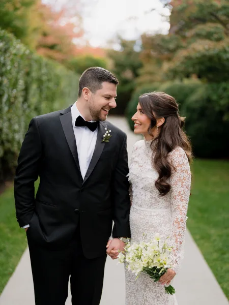 Couple portrait of a bride and groom walking hand in hand along a garden path, her long-sleeve lace bridal gown skimming the hedged walkway.