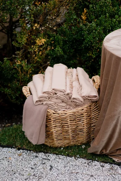Wedding blankets arranged in a guest blanket basket, neatly folded in a wicker basket beside garden shrubs along a gravel path.