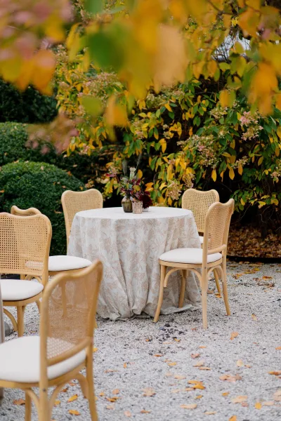 Cocktail hour table for outdoor cocktail seating, styled with a bud vase, amid autumn foliage and garden shrubs on gravel.
