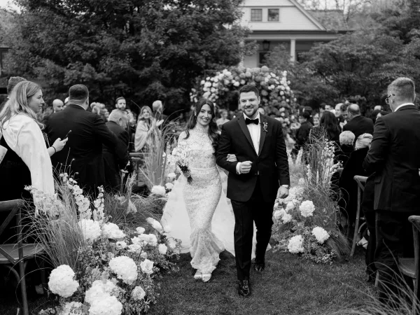 Wedding recessional with bride and groom walking down the aisle, smiling as guests applaud beneath a garden floral arch and shady trees.