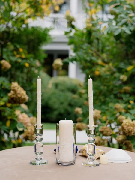 Unity candle setup for a candle lighting ceremony, with crystal candlesticks, framed by garden greenery and a house facade with porch columns.
