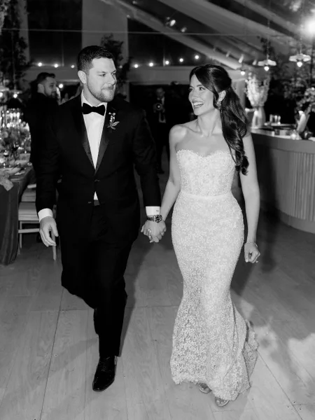 Reception entrance with a couple holding hands, the groom in a tuxedo beneath a tented ceiling glowing with string lights and reception tables