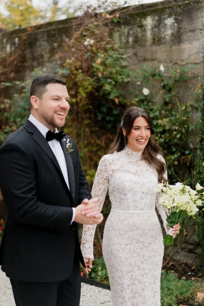Couple portrait of a bride and groom smiling, her lace bridal gown catching light as they stand by a vine-clad stone wall in the garden.