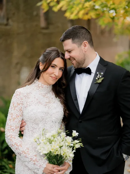 Couple portrait of bride and groom smiling as she holds a white bouquet before a stone wall framed by garden greenery and autumn leaves.