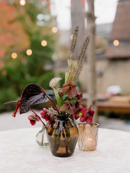 Cocktail table centerpiece with bud vase flowers arranged in an amber glass vase, under garden trees with string lights on an outdoor patio.