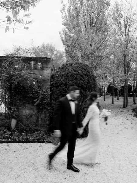 Couple portrait of the bride and groom walking, bride holding a bouquet along a tree-lined gravel path beside a stone wall.