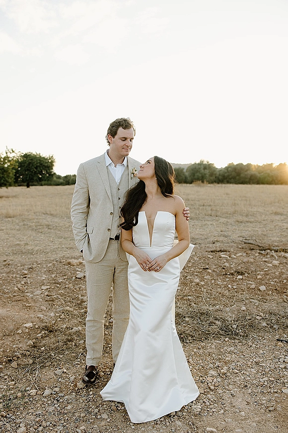 Couple portrait at sunset, outdoor wedding portrait of groom in beige suit holding bride in strapless plunge gown in a dry field