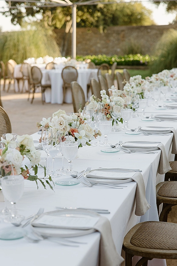 Reception tablescape with white linens, rose and orchid floral runner, silver flatware, and rattan chairs on a garden patio under a pergola