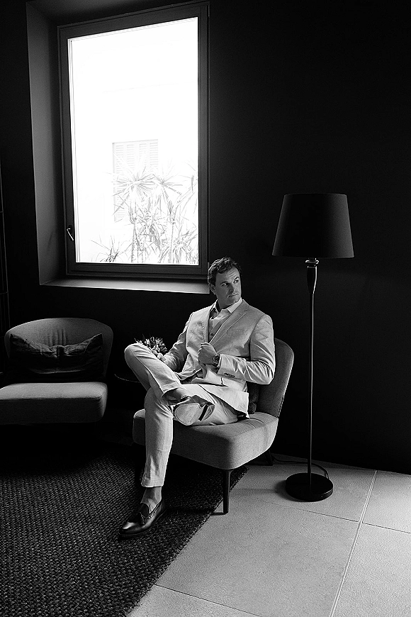 Groom portrait in a light suit seated in an armchair, boutonniere and wristwatch visible as he looks out a large window in a moody room