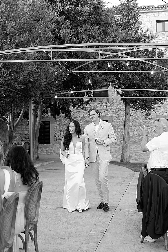 Wedding reception entrance as newlyweds grand entrance holding hands with champagne flutes under string lights in a stone courtyard with guests seated