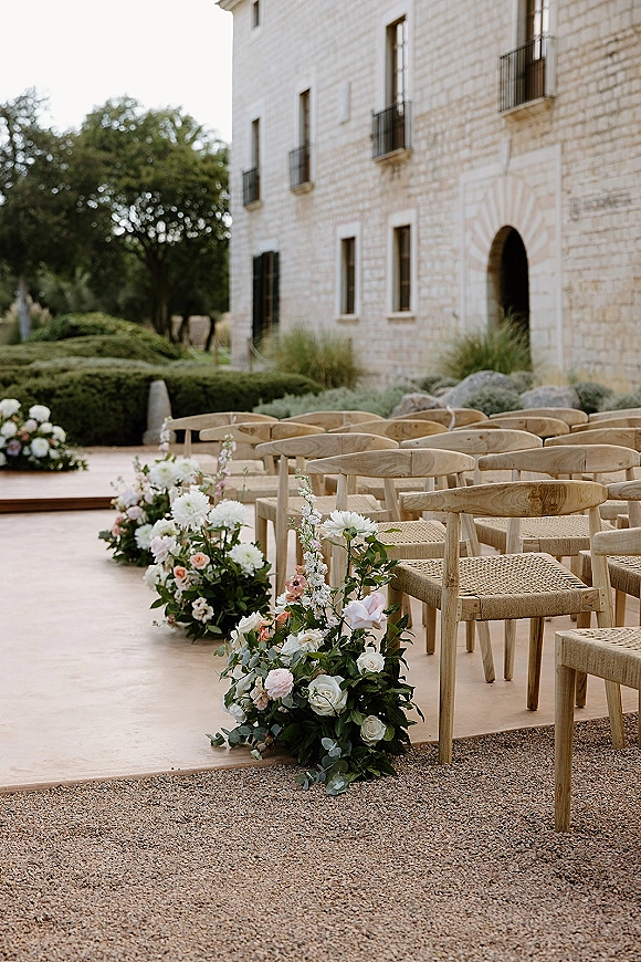 Ceremony aisle decor with woven outdoor ceremony chairs and low blush, white, and pink rose florals lining a wood platform by a stone archway