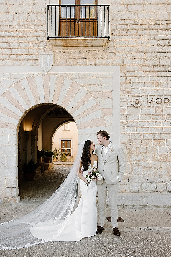 Couple portrait of bride and groom holding bouquet, gazing at each other beneath a stone archway, her cathedral veil trailing behind