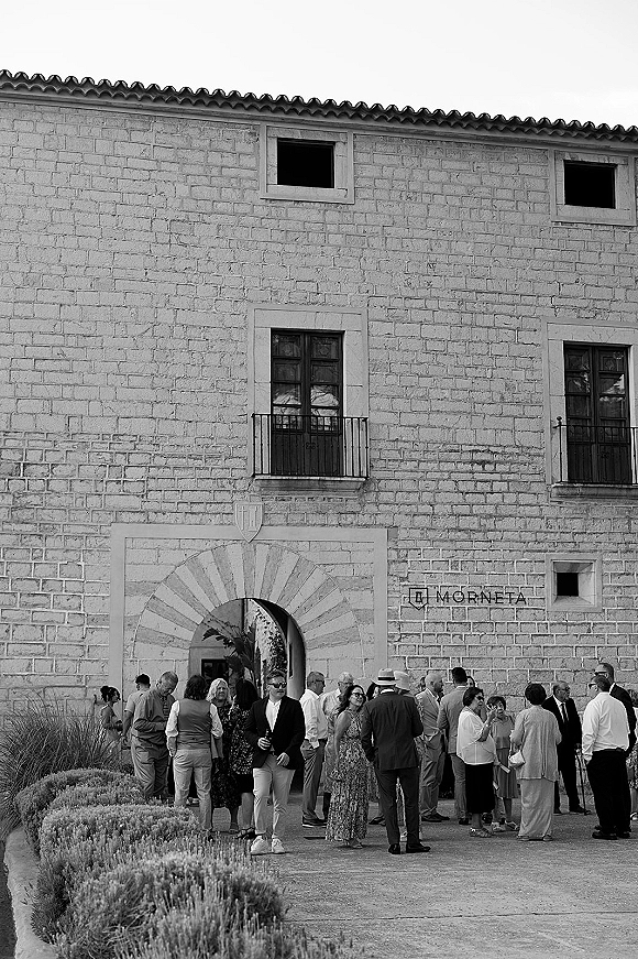 Wedding cocktail hour guests mingling in a courtyard, holding welcome drinks in suits and summer attire by a stone arched doorway facade