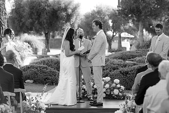 Wedding vows as bride reads into a microphone, holding hands with groom on a wooden platform in an outdoor garden ceremony, veil trailing