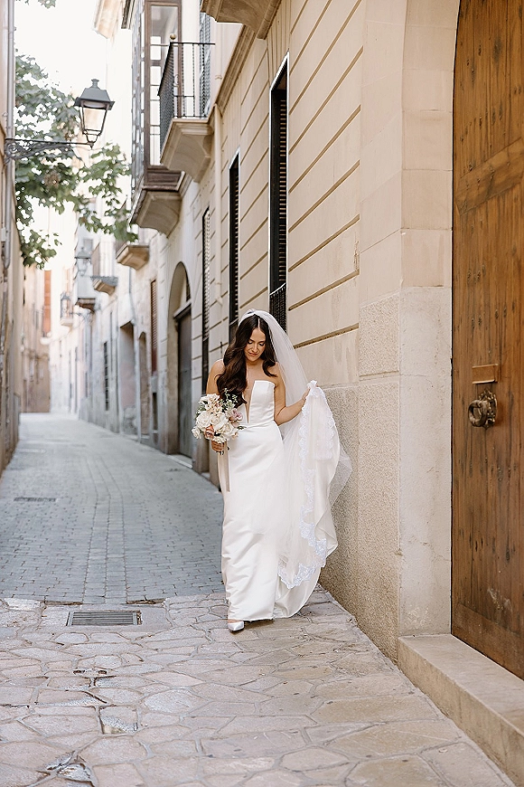 Bridal portrait of a bride in a strapless wedding dress with cathedral veil and bouquet walking a cobblestone old town street
