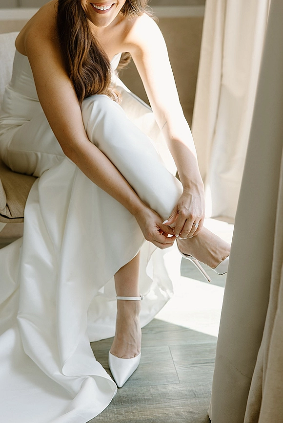 Bride getting ready as she sits on an upholstered chair putting on bridal shoes, engagement ring visible in soft window light