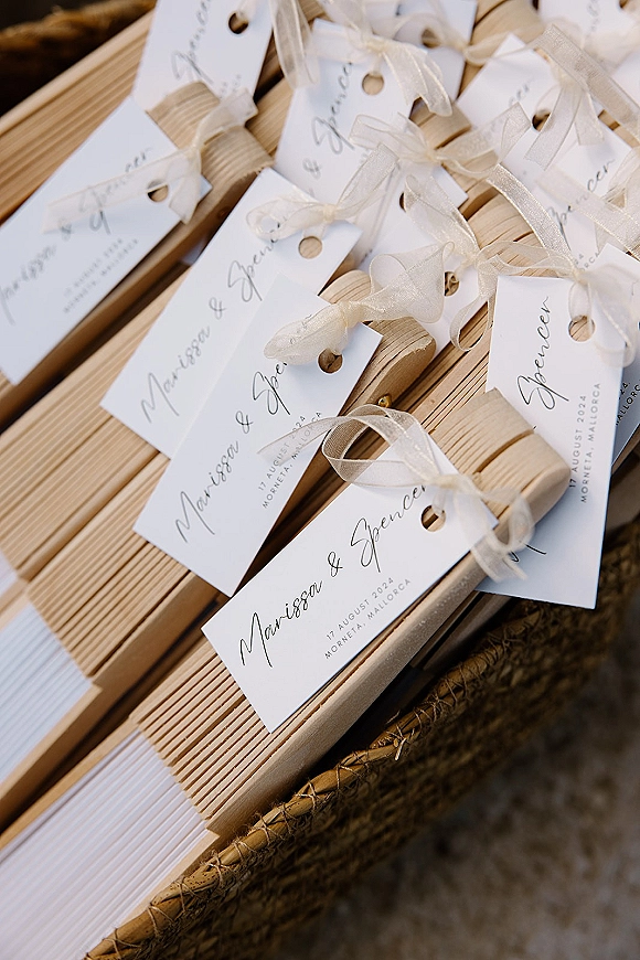 Wedding favors with personalized wedding favor tags in black script, ribbon bows, and wooden fans arranged in a woven basket on slats