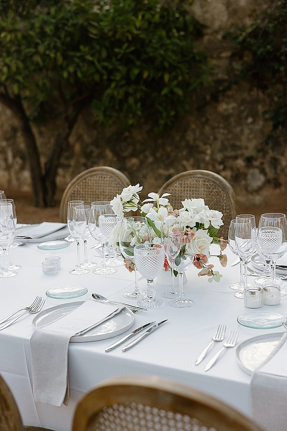 Reception tablescape with white tablecloth, linen napkins, floral centerpiece, candles and textured goblets, set outdoors by a stone wall
