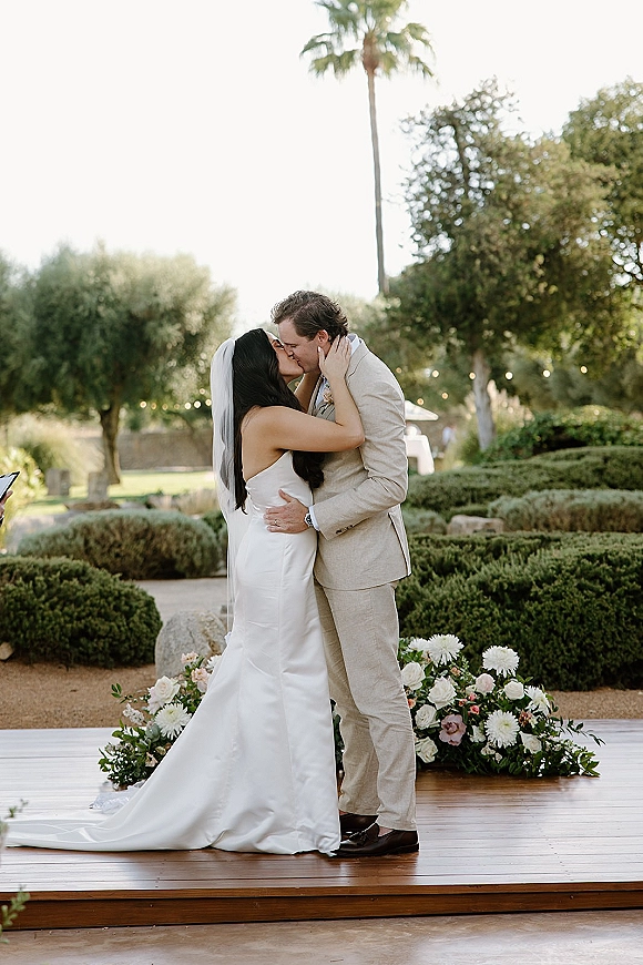 Wedding kiss portrait of bride and groom kissing on a wooden platform, her long veil flowing at a garden ceremony with string lights