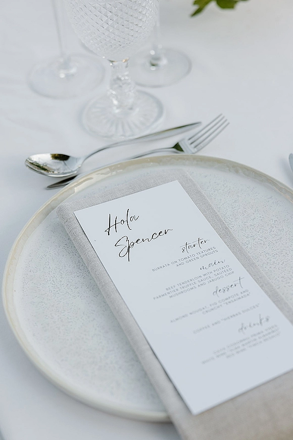 Wedding place setting with wedding menu card and calligraphy place card on a linen napkin, ceramic plate, silver cutlery, and goblets on white tablecloth with greenery