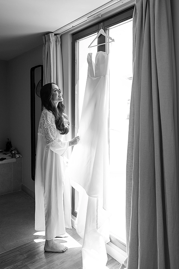 Bride getting ready in a bridal robe with lace sleeves, gazing at a wedding dress on hanger by the window in a bedroom mirror reflection