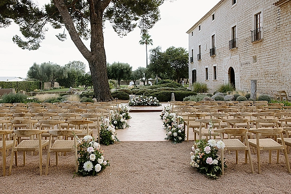 Outdoor ceremony setup with wood chairs and aisle floral arrangements lining a gravel aisle in a garden courtyard by a stone building backdrop
