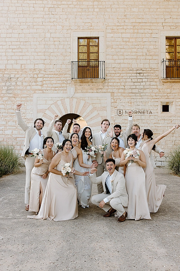 Wedding party portrait of bride with bridesmaids and groom with groomsmen holding bouquets, laughing in a gravel courtyard by a stone building facade