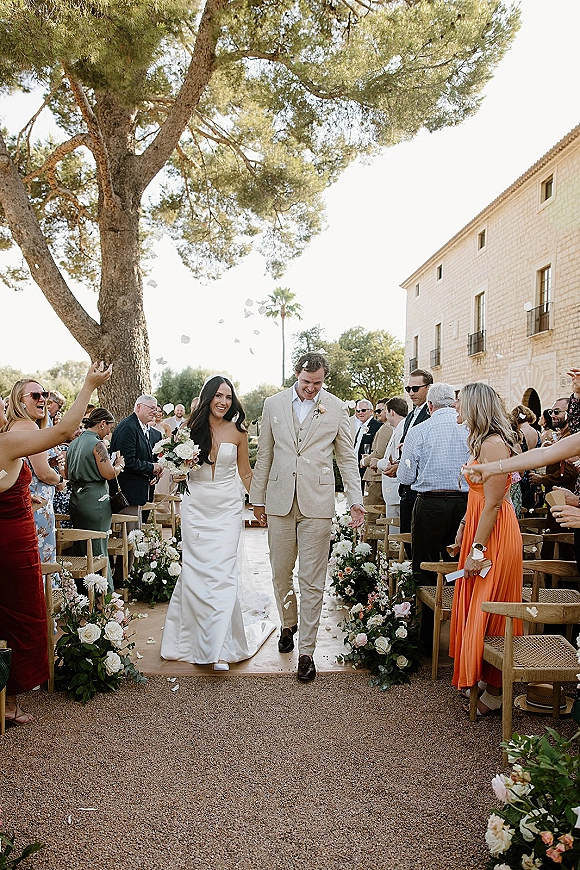 Wedding recessional as bride and groom walk up the outdoor aisle, bouquet in hand, guests cheering beside flower petals and chairs under a large tree