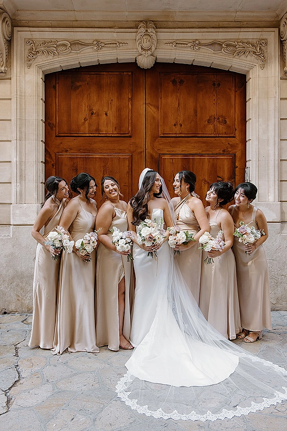 Bride with bridesmaids in a bridesmaids group photo, all holding pastel bouquets in champagne dresses by wooden doors and stone archway