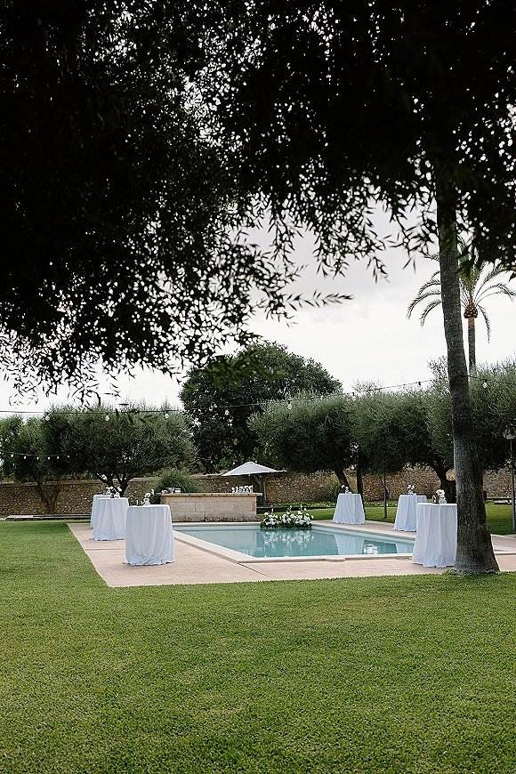 Poolside cocktail hour setup with white linen cocktail tables and floral centerpieces under string lights beside a swimming pool and palm tree
