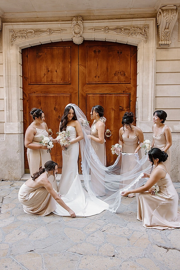 Bride with bridesmaids as they fix her cathedral veil and strapless gown, holding blush-and-white bouquets before wooden doors and stone archway