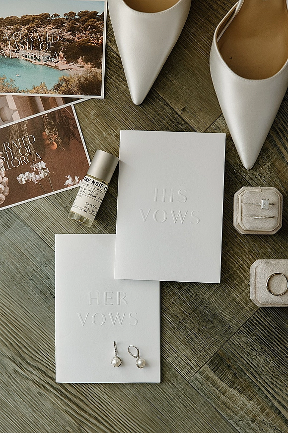 Vow book flatlay with his and her vow books, white pointed-toe heels, rings, pearl drop earrings, perfume, and postcards on wood floor
